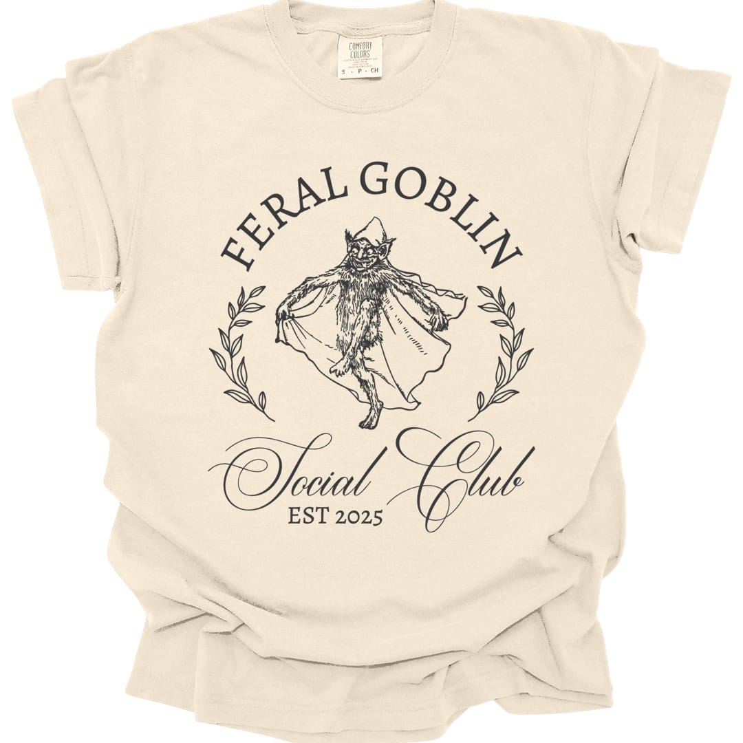 Ivory t-shirt. Reads "Feral Goblin" curving across the top and "Social Club" in script across the bottom, with "Est 2025" beneath. In the middle a long-haired cat stands upright in a flowing cloak inside a thin laurel wreath. Delicate gray line work throughout.