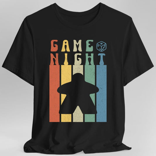 Black t-shirt. Reads "Game Night" in chunky letters across the top, each letter in a different retro color. Below, a large gray meeple stands in front of five tall vertical stripes in orange, gold, cream, green and blue. A small die sits by the top right.