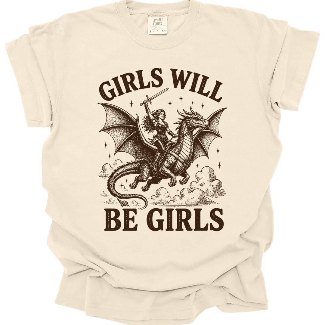 Ivory t-shirt. The design reads Girls Will arched across the top and Be Girls across the bottom in chunky distressed capitals. In between, a woman adventurer in leather armor with long flowing hair rides a large winged dragon through the clouds, holding a sword raised high above her head. Dark brown woodcut lines with small stars scattered around.