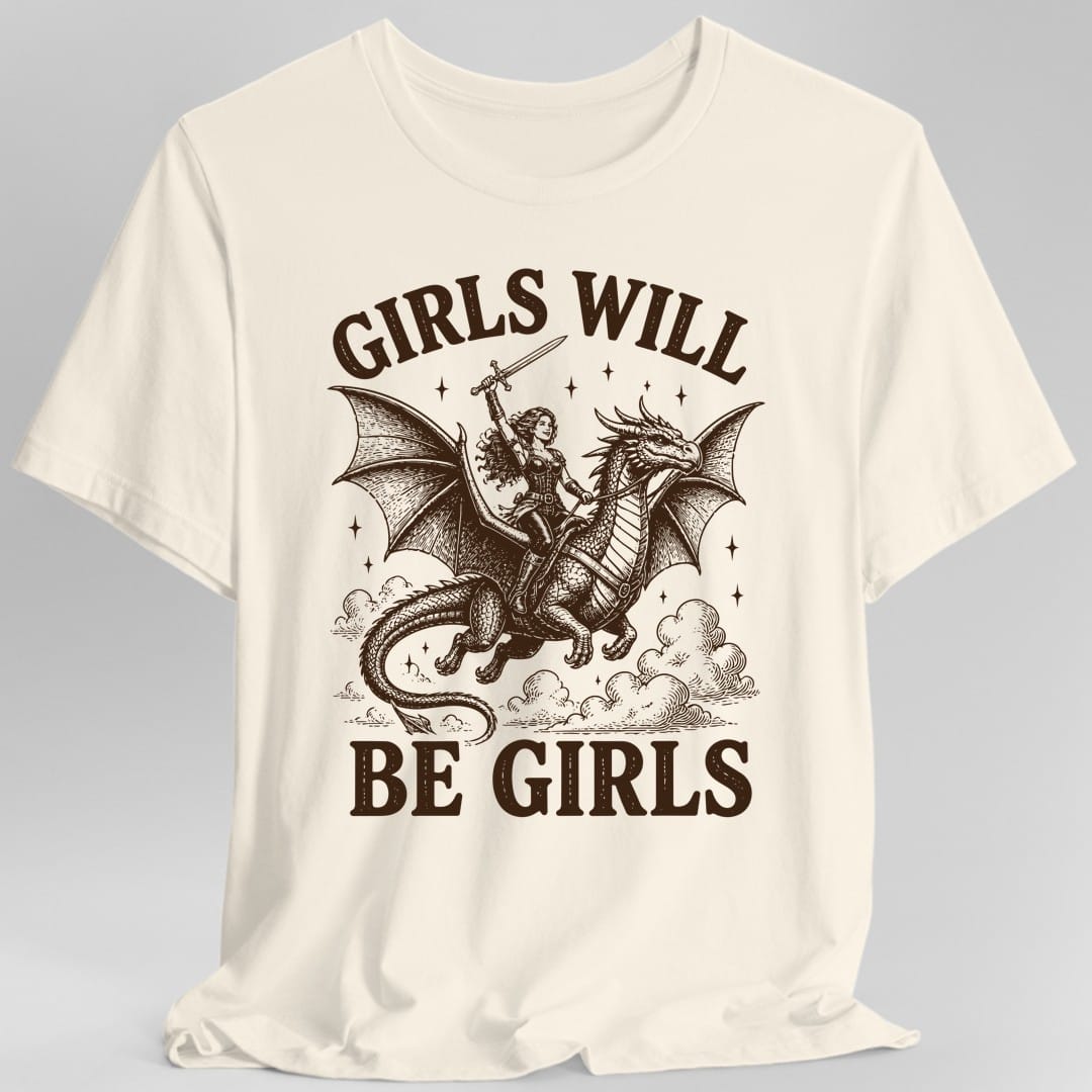 Natural t-shirt. The design reads Girls Will arched across the top and Be Girls across the bottom in chunky distressed capitals. In between, a woman adventurer in leather armor with long flowing hair rides a large winged dragon through the clouds, holding a sword raised high above her head. Dark brown woodcut lines with small stars scattered around.