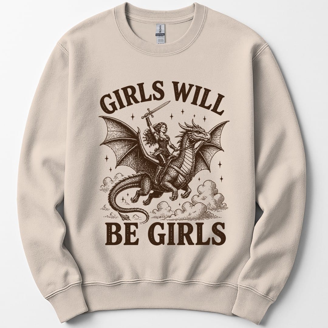 Sand sweatshirt. The design reads Girls Will arched across the top and Be Girls across the bottom in chunky distressed capitals. In between, a woman adventurer in leather armor with long flowing hair rides a large winged dragon through the clouds, holding a sword raised high above her head. Dark brown woodcut lines with small stars scattered around.