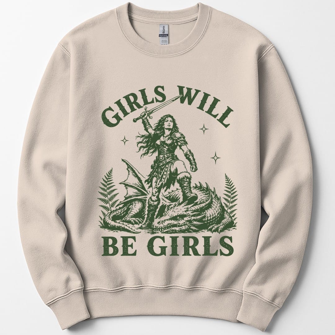 Sand sweatshirt. Reads "Girls will" across the top and "Be girls" across the bottom in bold green capitals. Between them a woman in armor stands over a slain dragon, sword raised above her head, with ferns and sparkles around her. Engraved linework, all in one forest green.