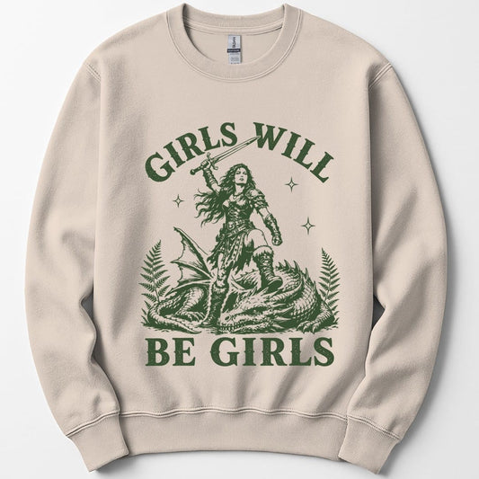 Sand sweatshirt. Reads "Girls will" across the top and "Be girls" across the bottom in bold green capitals. Between them a woman in armor stands over a slain dragon, sword raised above her head, with ferns and sparkles around her. Engraved linework, all in one forest green.