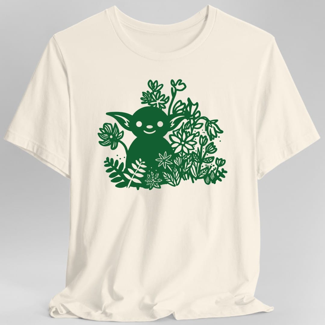 Natural t-shirt. A single color green cut paper style illustration. A round little goblin with wide pointed ears, dark eyes and a small smile sits up to its chest in a thicket of leaves, ferns and wildflowers that surround it on all sides and rise above its head. No words.