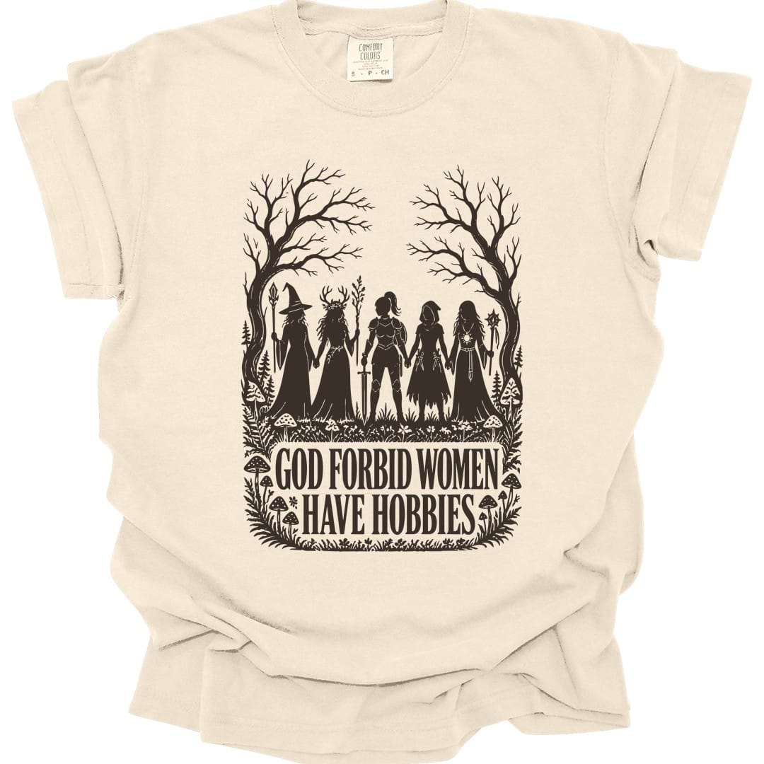 Ivory t-shirt. The design reads "God Forbid Women Have Hobbies" in bold serif capitals across two lines at the bottom. Above the words five women adventurers stand in silhouette holding hands in a row, a wizard with a staff, a druid crowned with antlers, a knight in armor with a sword, a hooded ranger with daggers, and a sorcerer with an orb topped staff. Bare trees arch overhead and mushrooms and ferns crowd the ground. Cut paper silhouette art in one dark brown ink.