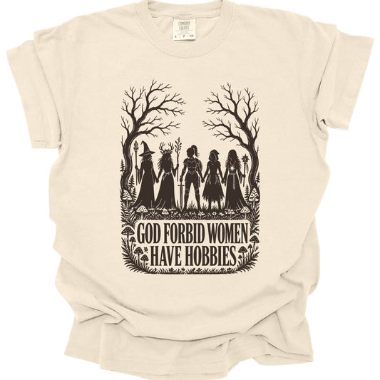 Ivory t-shirt. The design reads "God Forbid Women Have Hobbies" in bold serif capitals across two lines at the bottom. Above the words five women adventurers stand in silhouette holding hands in a row, a wizard with a staff, a druid crowned with antlers, a knight in armor with a sword, a hooded ranger with daggers, and a sorcerer with an orb topped staff. Bare trees arch overhead and mushrooms and ferns crowd the ground. Cut paper silhouette art in one dark brown ink.