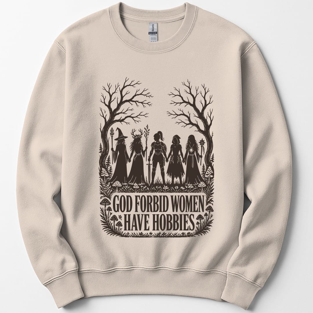 Sand sweatshirt. The design reads "God Forbid Women Have Hobbies" in bold serif capitals across two lines at the bottom. Above the words five women adventurers stand in silhouette holding hands in a row, a wizard with a staff, a druid crowned with antlers, a knight in armor with a sword, a hooded ranger with daggers, and a sorcerer with an orb topped staff. Bare trees arch overhead and mushrooms and ferns crowd the ground. Cut paper silhouette art in one dark brown ink.
