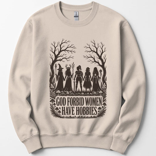 Sand sweatshirt. The design reads "God Forbid Women Have Hobbies" in bold serif capitals across two lines at the bottom. Above the words five women adventurers stand in silhouette holding hands in a row, a wizard with a staff, a druid crowned with antlers, a knight in armor with a sword, a hooded ranger with daggers, and a sorcerer with an orb topped staff. Bare trees arch overhead and mushrooms and ferns crowd the ground. Cut paper silhouette art in one dark brown ink.