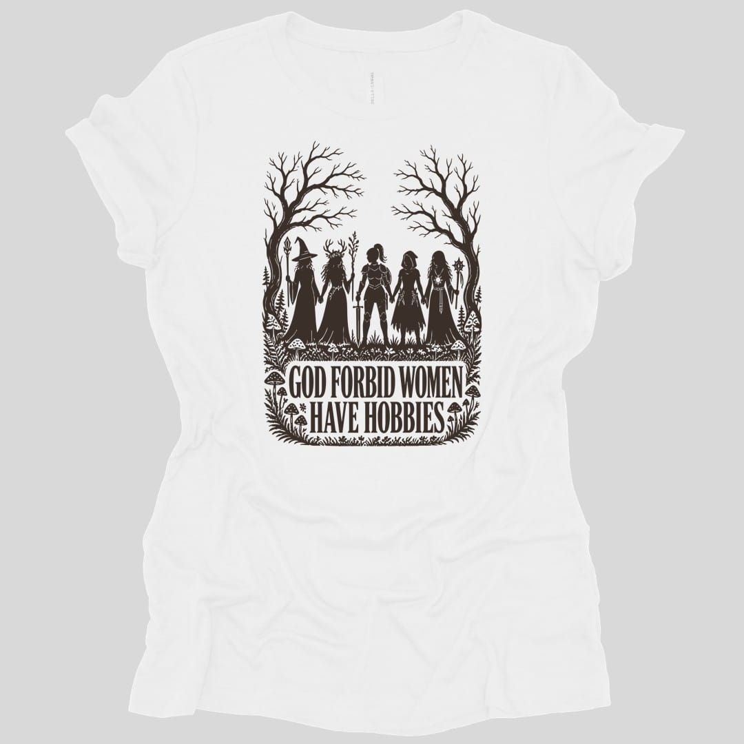 White women's t-shirt. The design reads "God Forbid Women Have Hobbies" in bold serif capitals across two lines at the bottom. Above the words five women adventurers stand in silhouette holding hands in a row, a wizard with a staff, a druid crowned with antlers, a knight in armor with a sword, a hooded ranger with daggers, and a sorcerer with an orb topped staff. Bare trees arch overhead and mushrooms and ferns crowd the ground. Cut paper silhouette art in one dark brown ink.