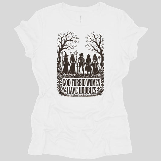 White women's t-shirt. The design reads "God Forbid Women Have Hobbies" in bold serif capitals across two lines at the bottom. Above the words five women adventurers stand in silhouette holding hands in a row, a wizard with a staff, a druid crowned with antlers, a knight in armor with a sword, a hooded ranger with daggers, and a sorcerer with an orb topped staff. Bare trees arch overhead and mushrooms and ferns crowd the ground. Cut paper silhouette art in one dark brown ink.