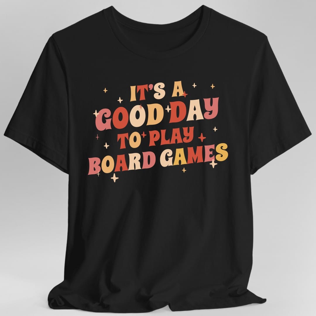 Black t-shirt. Reads "It's a good day to play board games" across four lines in fat seventies bubble lettering, each word in coral, cream or gold, tilted slightly. Small four-pointed sparkles are scattered between the words. All type.