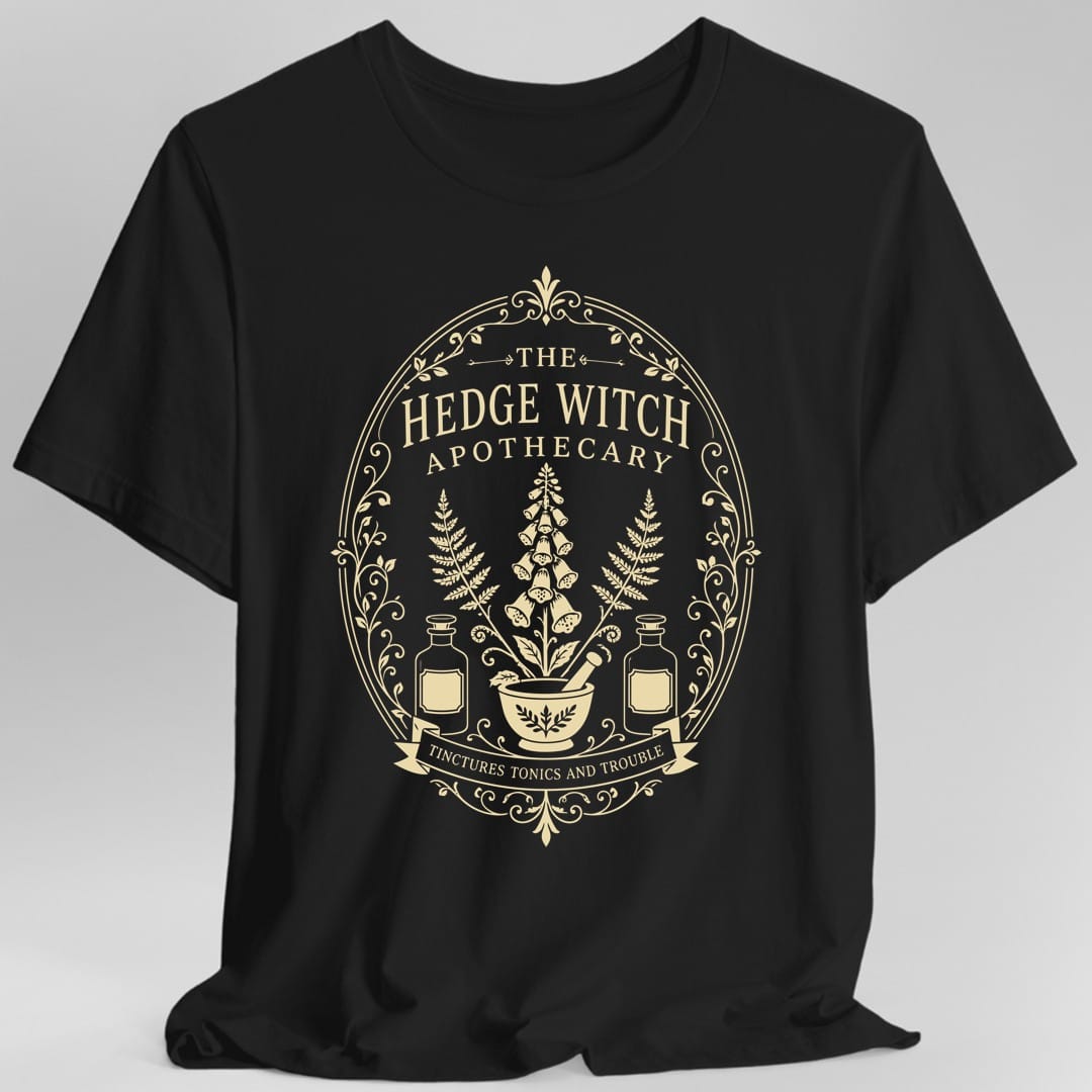 Black t-shirt. The design is an oval apothecary label in cream line work. THE sits at the top between small arrows, then HEDGE WITCH in tall capitals and APOTHECARY spaced below it. Inside the frame a tall foxglove spike rises between two ferns, above a mortar and pestle flanked by two stoppered tincture bottles. A ribbon banner across the bottom reads TINCTURES TONICS AND TROUBLE, with scrollwork and vines around the border.