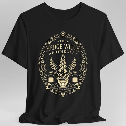 Black t-shirt. The design is an oval apothecary label in cream line work. THE sits at the top between small arrows, then HEDGE WITCH in tall capitals and APOTHECARY spaced below it. Inside the frame a tall foxglove spike rises between two ferns, above a mortar and pestle flanked by two stoppered tincture bottles. A ribbon banner across the bottom reads TINCTURES TONICS AND TROUBLE, with scrollwork and vines around the border.