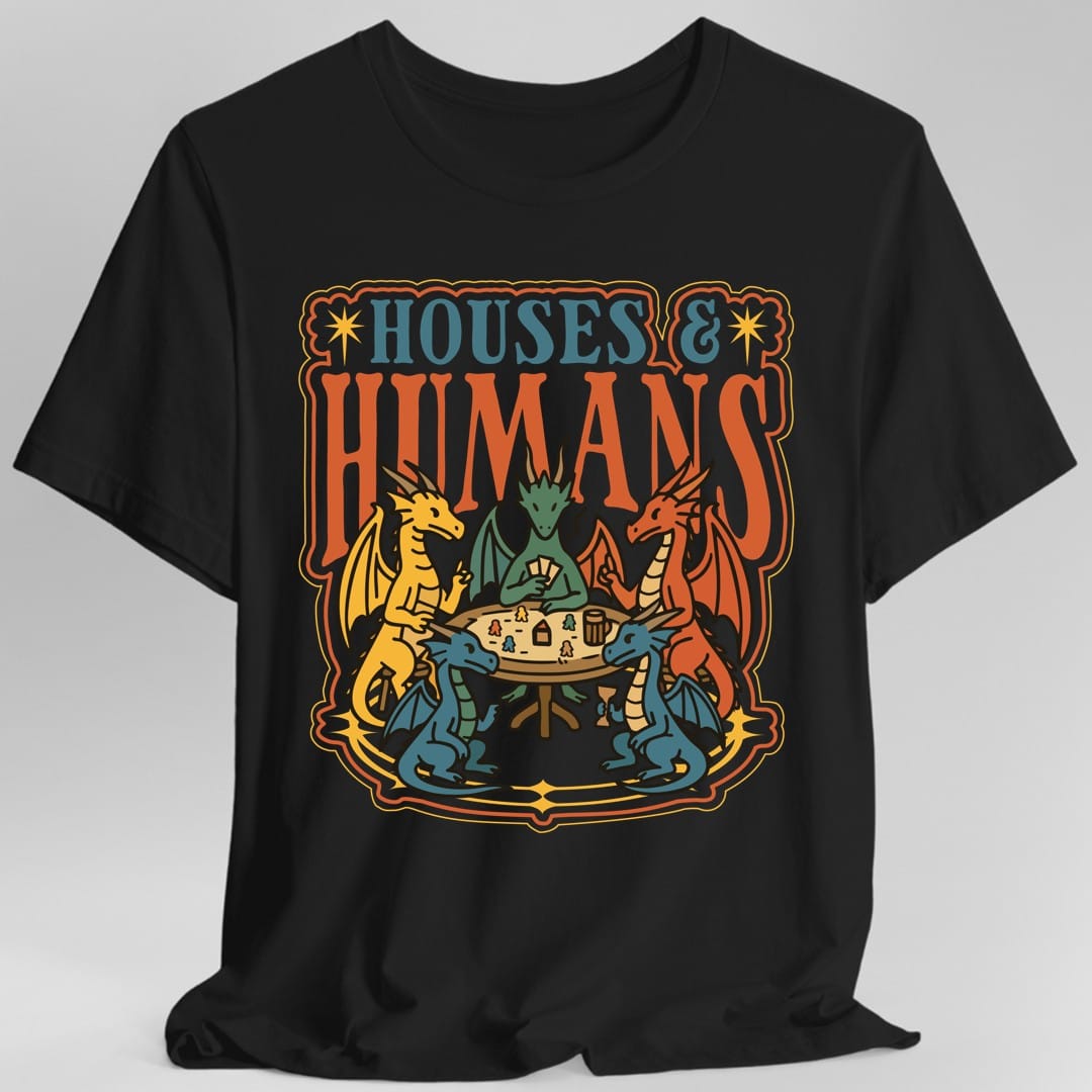 Black t-shirt. The design reads "Houses &" in blue serif capitals and "Humans" in tall orange capitals beneath it. Below the words five dragons in yellow, green, red and blue sit around a round wooden table playing a board game, one holding a fan of cards, with tiny human game pieces, little houses, a tankard and a goblet on the table between them. An ornate orange and gold border frames the whole scene. Flat bold outlined illustration.