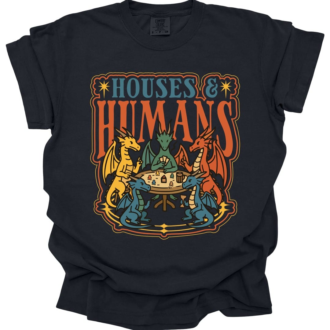 Black t-shirt. The design reads "Houses &" in blue serif capitals and "Humans" in tall orange capitals beneath it. Below the words five dragons in yellow, green, red and blue sit around a round wooden table playing a board game, one holding a fan of cards, with tiny human game pieces, little houses, a tankard and a goblet on the table between them. An ornate orange and gold border frames the whole scene. Flat bold outlined illustration.