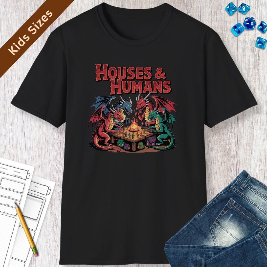 Black kids t-shirt laid flat with dice and jeans. The design reads "Houses and Humans" in tall red horror-comic lettering across the top. Below it, four dragons in red, blue, green and crimson sit around a wooden table like players at game night, leaning in over a small campfire in the middle. Tiny human figures in blue, green and red stand scattered across the tabletop, and little cottages and tents sit on the ground underneath. Bold outlined cartoon art in red, teal, blue and gold.