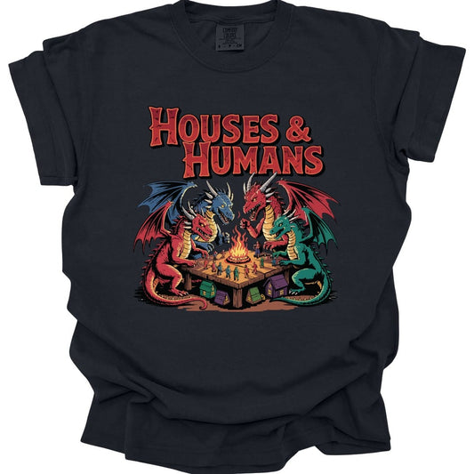 Black t-shirt. The design reads "Houses and Humans" in tall red horror-comic lettering across the top. Below it, four dragons in red, blue, green and crimson sit around a wooden table like players at game night, leaning in over a small campfire in the middle. Tiny human figures in blue, green and red stand scattered across the tabletop, and little cottages and tents sit on the ground underneath. Bold outlined cartoon art in red, teal, blue and gold.