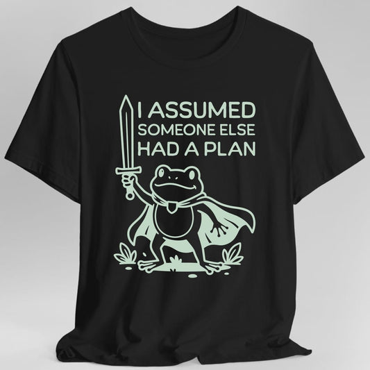 Black t-shirt. Reads "I Assumed Someone Else Had A Plan" across the top in three lines of rounded capitals. Below it a frog stands upright in a flowing cape, holding a sword straight up in one webbed hand, surrounded by reeds and lily pads. Simple single color outline drawing.