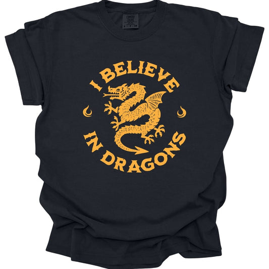 Black t-shirt. Reads "I believe" curving across the top and "In dragons" across the bottom in capitals, forming a circle. Inside sits a long serpentine dragon coiled around itself with a crescent moon either side. All in one color, golden yellow on dark shirts and deep red on light ones.