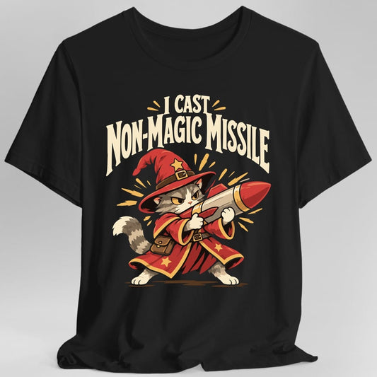 Black t-shirt. The design reads I CAST in cream capitals at the top and NON-MAGIC MISSILE in large arched cream capitals below it, with yellow spark marks around the words. Underneath, a grey and white cat with a stern expression stands in a red wizard hat and a long red robe trimmed in gold stars, a brown satchel at its belt, hoisting a large red tipped rocket across its body like a weapon.