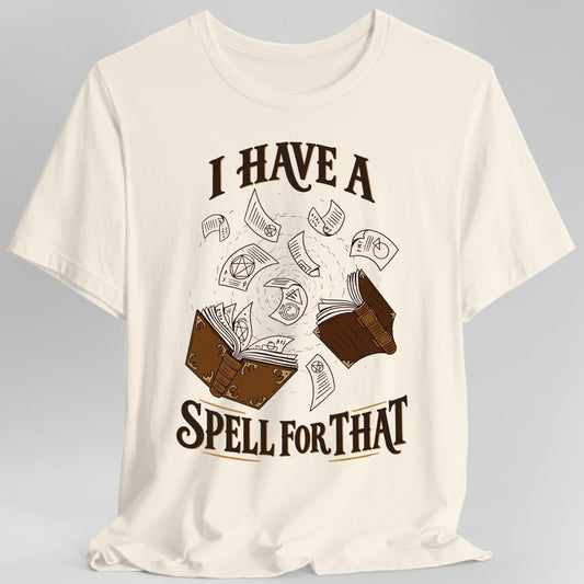 Natural t-shirt. The design reads I HAVE A in dark brown capitals arched across the top and SPELL FOR THAT in large capitals along the bottom, underlined with thin gold strokes. Between them two leather bound spellbooks with brass clasps lie open facing each other, and loose cream pages marked with pentacles, runes and diagrams swirl upward in a spiral between them.