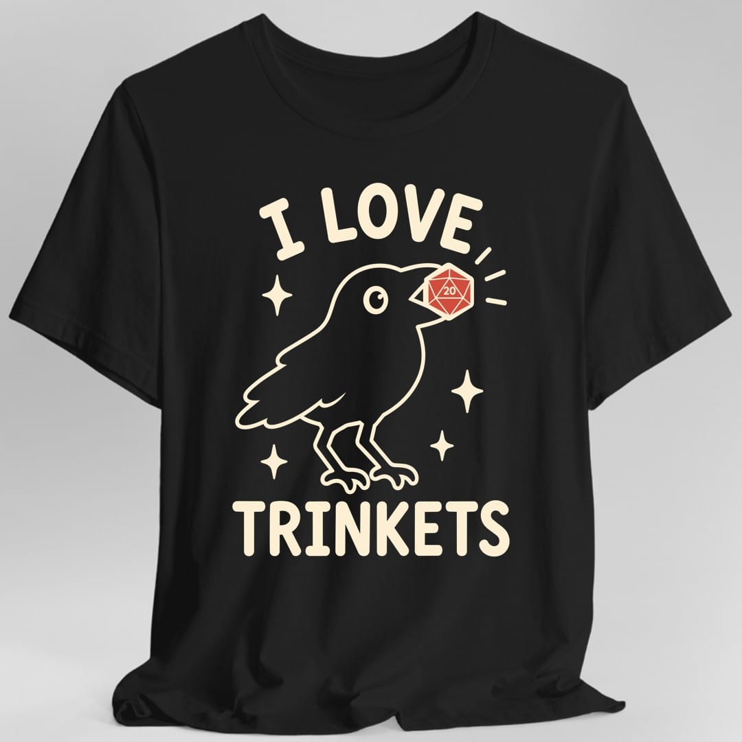 Black t-shirt. The design reads I LOVE at the top and TRINKETS at the bottom in rounded cream capitals. Between them a crow drawn in simple outline holds a red twenty sided die in its beak, the number 20 showing on the face. Small four pointed sparkles float around the bird. Clean flat line art in cream and red.