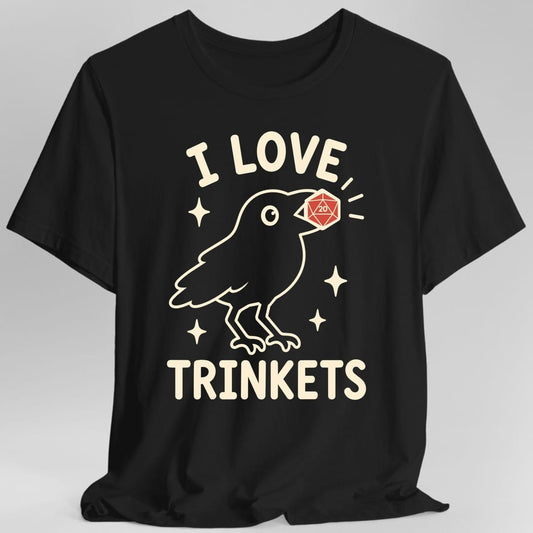 Black t-shirt. The design reads I LOVE at the top and TRINKETS at the bottom in rounded cream capitals. Between them a crow drawn in simple outline holds a red twenty sided die in its beak, the number 20 showing on the face. Small four pointed sparkles float around the bird. Clean flat line art in cream and red.