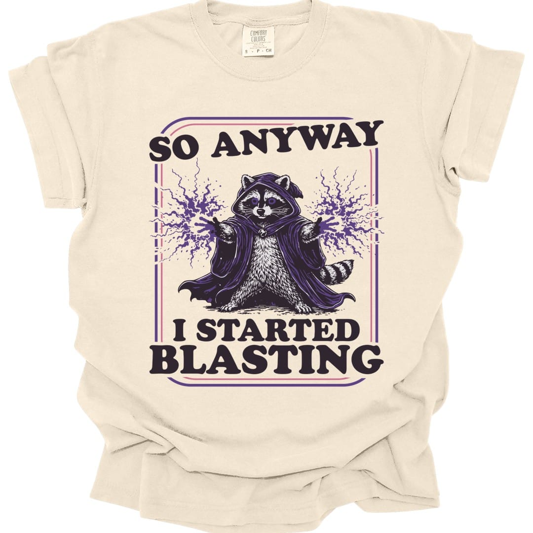 Ivory t-shirt. Reads "So anyway" across the top in dark capitals and "I started blasting" across the bottom. Between them a raccoon sits in a dark hooded cloak with both paws raised, purple magic crackling out from each one. Engraved linework in near-black and purple, no frame.