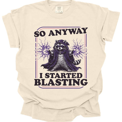 Ivory t-shirt. Reads "So anyway" across the top in dark capitals and "I started blasting" across the bottom. Between them a raccoon sits in a dark hooded cloak with both paws raised, purple magic crackling out from each one. Engraved linework in near-black and purple, no frame.