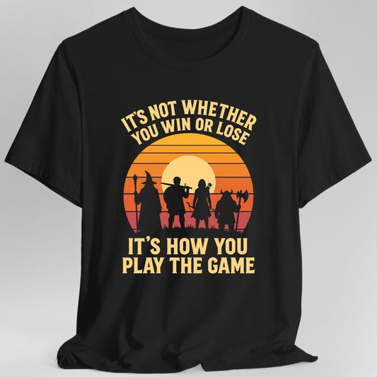 Black t-shirt. The design reads "It's not whether you win or lose" curved across the top and "It's how you play the game" in two big lines at the bottom, in pale gold letters. Between them, four adventurers stand in silhouette on grass: a wizard with a staff, a fighter shouldering a sword, an archer with a quiver, and a horned barbarian with an axe. Behind them is a pale moon inside a striped sunset going from orange down to deep red.
