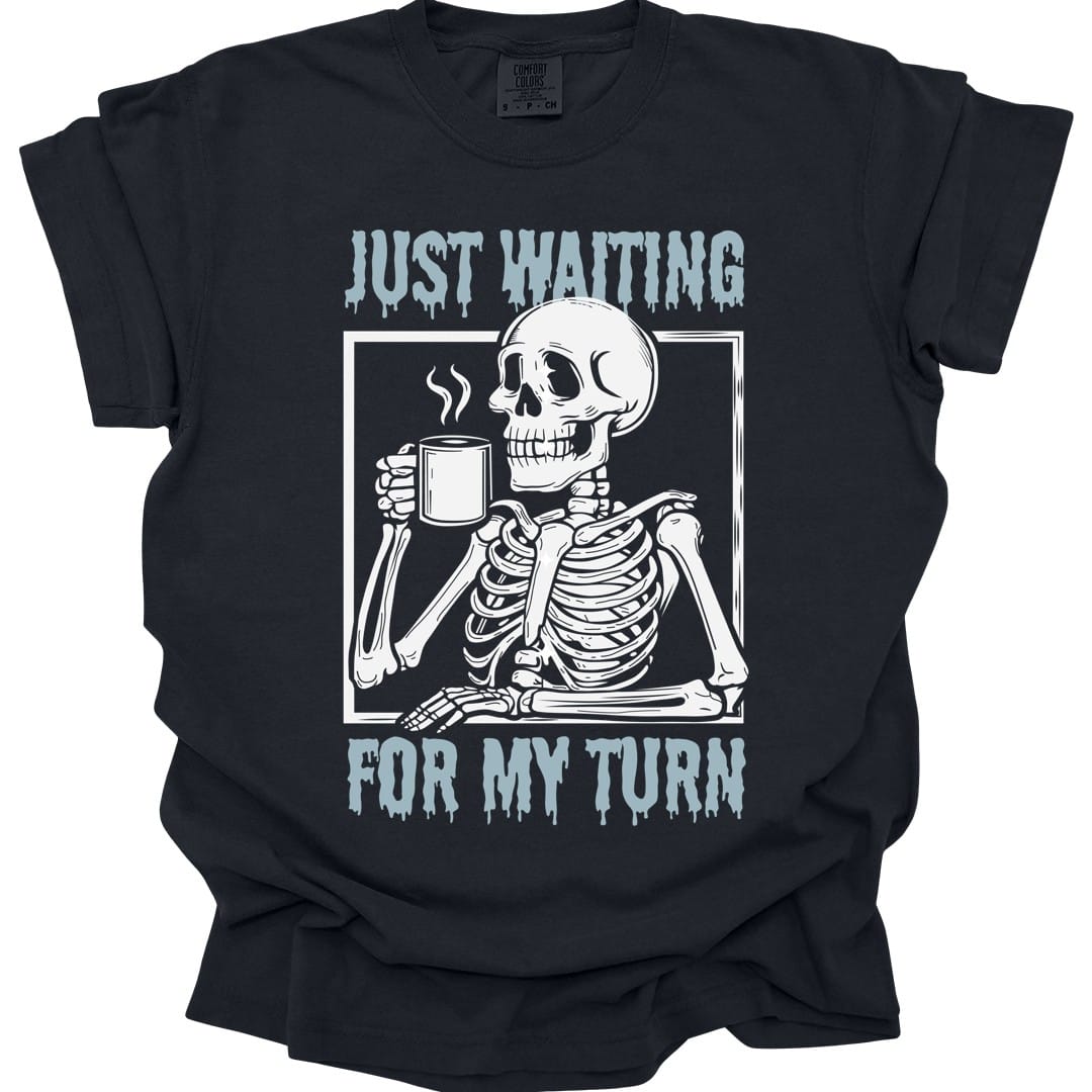 Black t-shirt. Reads "Just waiting" across the top and "For my turn" across the bottom in pale blue capitals with paint dripping from them. Between them a skeleton sits at a table inside a thin frame, holding a steaming mug, elbow propped up.