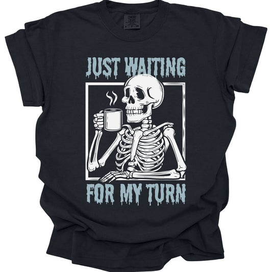 Black t-shirt. Reads "Just waiting" across the top and "For my turn" across the bottom in pale blue capitals with paint dripping from them. Between them a skeleton sits at a table inside a thin frame, holding a steaming mug, elbow propped up.