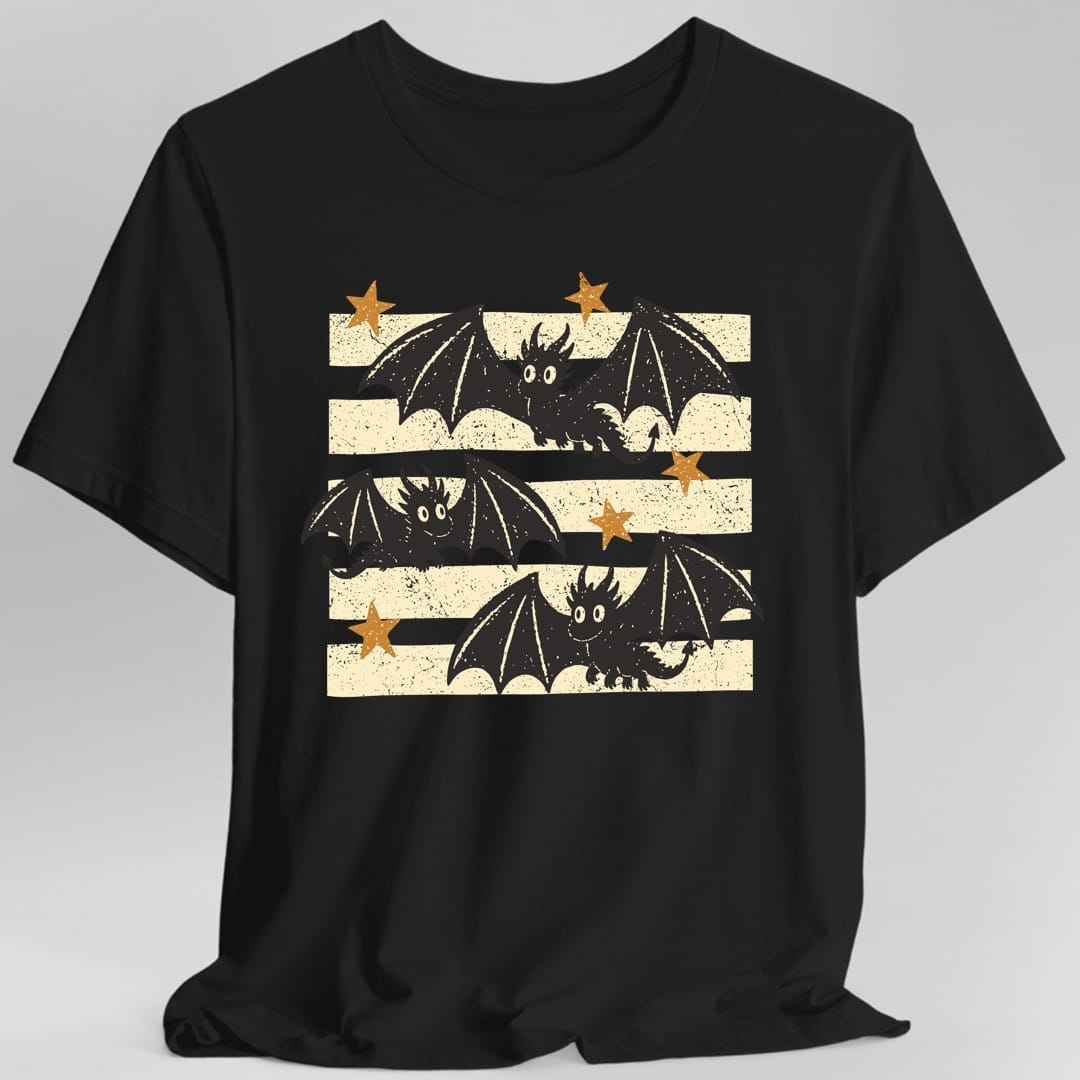 Black t-shirt. The design shows three small black dragons with round white eyes, tufted horns and wide spread bat wings, flying one above another across a block of wide cream colored stripes with a rough, speckled printed texture. Their wings and arrow tipped tails reach out past the edges of the stripes. Five gold stars sit among them. Hand printed woodblock style, with no words.
