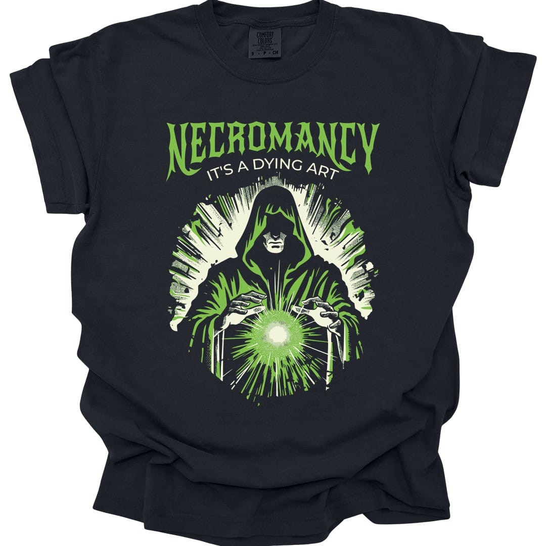 Black t-shirt. Reads "Necromancy" across the top in green capitals with "It's a dying art" beneath. Below, a hooded figure stands with both hands raised over a glowing ball of green light, its face lost in shadow, with green energy radiating out behind it.
