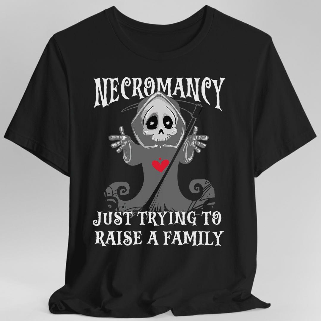 Black t-shirt. Reads "Necromancy" across the top and "Just trying to raise a family" across the bottom in white spooky capitals. Between them a small cartoon grim reaper in a gray hood holds up both bony hands with a friendly wide-eyed skull face and a red heart on its chest, a scythe leaning behind it. Cute rather than scary. Greys and white with the one red heart.