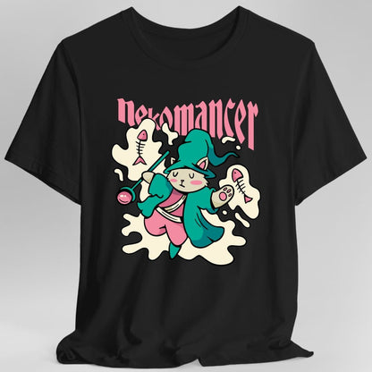 Black t-shirt. Reads "Nekomancer" across the top in pink blackletter. Below, a white cat in a teal hooded robe holds up a staff topped with a pink orb, standing among floating fish skeletons and a pale cloud of magic. Soft cartoon style in teal, pink and cream.