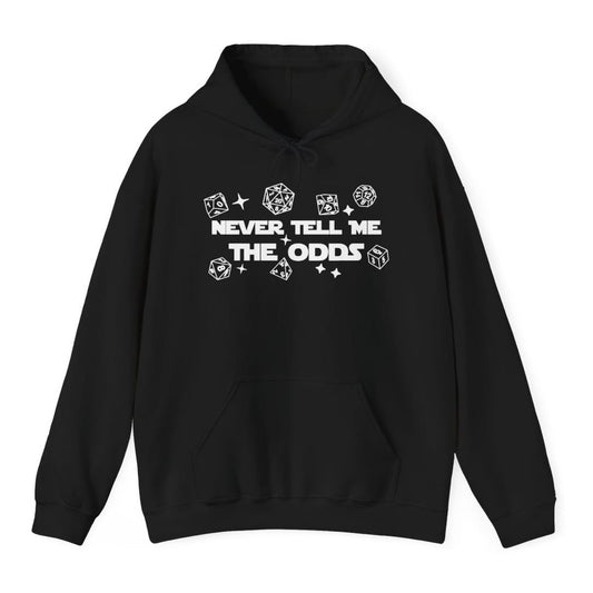 Black hoodie. Reads "Never tell me" over "The odds" in bold white capitals across the middle. Polyhedral dice of every shape float loose around the words, drawn as simple white outlines, with a few four-pointed sparkles between them.