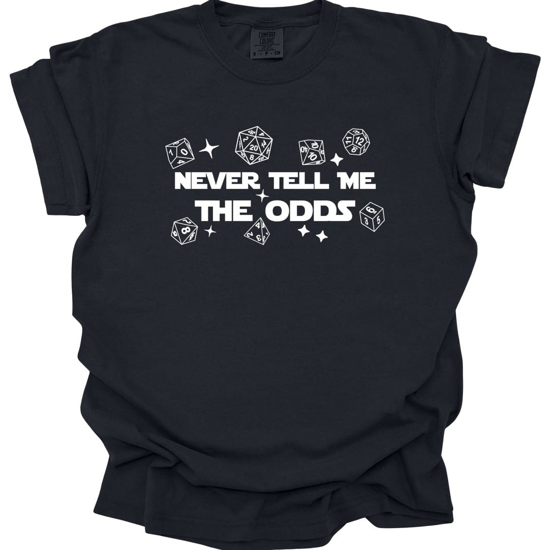 Black t-shirt. Reads "Never tell me" over "The odds" in bold white capitals across the middle. Polyhedral dice of every shape float loose around the words, drawn as simple white outlines, with a few four-pointed sparkles between them.