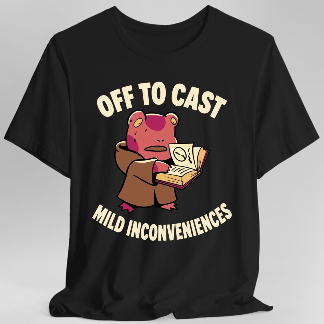 Black t-shirt. The design reads "OFF TO CAST" arched above and "MILD INCONVENIENCES" curved below, in cream letters. Between them a chunky pink frog in a brown wizard robe holds an open spellbook in both hands, beads of sweat on its brow and its mouth drawn as a flat worried line. One page of the book shows a scribbled diagram.