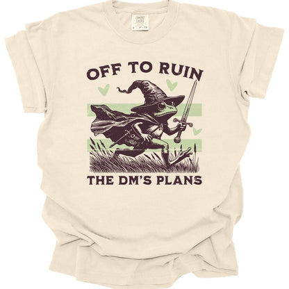 Ivory t-shirt. Reads "Off to ruin" across the top and "the DM's plans" across the bottom in chunky deep plum capitals. Between them a frog in a floppy pointed wizard hat and cape dashes to the right through reeds and cattails, holding a sword aloft. Two pale sage stripes run behind it and three sage hearts float around it. Retro woodcut style in deep plum and pale sage green.