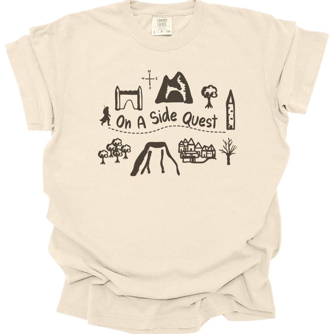 Ivory t-shirt. A hand-drawn map layout in brown line work: a stone archway, a mountain with a cave, trees, a tower, a village, a volcano and a small figure, with a dotted trail winding between them. "On a side quest" is written across the middle of the trail.