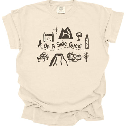 Ivory t-shirt. A hand-drawn map layout in brown line work: a stone archway, a mountain with a cave, trees, a tower, a village, a volcano and a small figure, with a dotted trail winding between them. "On a side quest" is written across the middle of the trail.