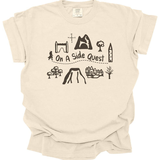 Ivory t-shirt. A hand-drawn map layout in brown line work: a stone archway, a mountain with a cave, trees, a tower, a village, a volcano and a small figure, with a dotted trail winding between them. "On a side quest" is written across the middle of the trail.