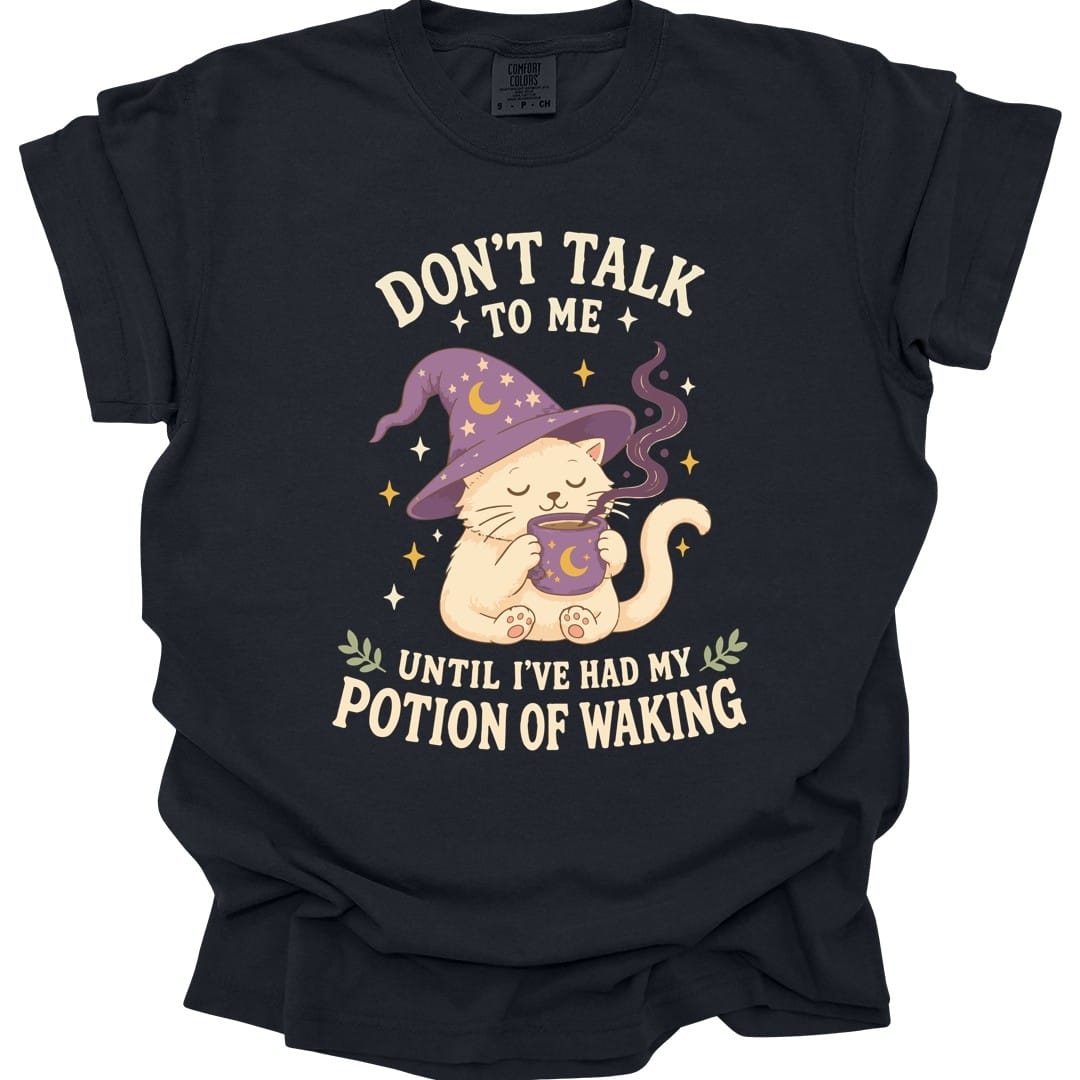 Black t-shirt. Reads "Don't talk" across the top with "to me" beneath, and "Until I've had my" and "Potion of waking" across the bottom in cream capitals. Between them a cream cat in a purple starry wizard hat holds a steaming mug in both paws, eyes closed.