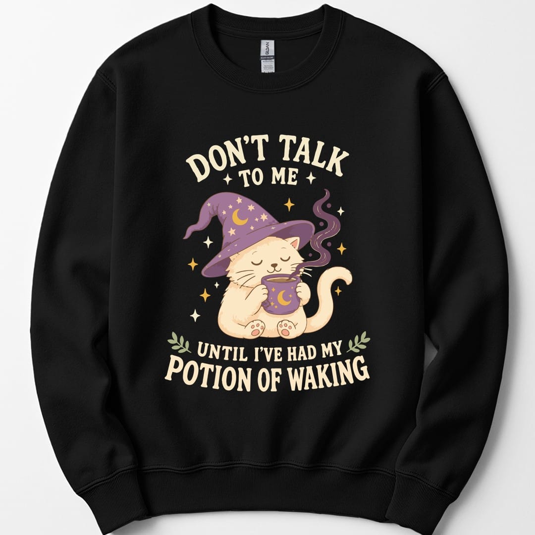 Black sweatshirt. Reads "Don't talk" across the top with "to me" beneath, and "Until I've had my" and "Potion of waking" across the bottom in cream capitals. Between them a cream cat in a purple starry wizard hat holds a steaming mug in both paws, eyes closed.