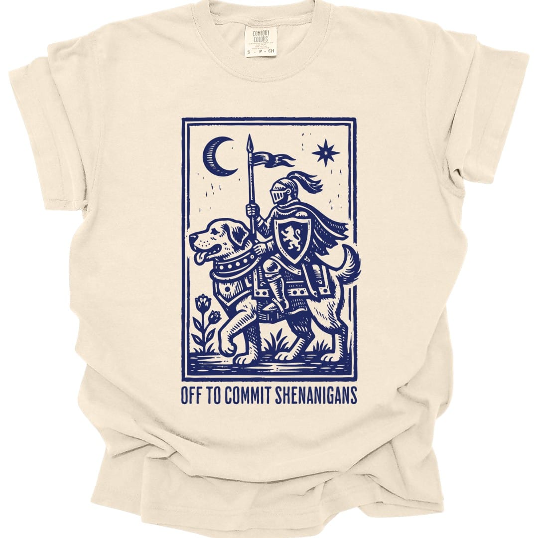 Ivory t-shirt. A knight in full armor rides a large hound like a horse, carrying a lance with a pennant, under a crescent moon and a star, all inside a thin ruled border. Underneath, "Off to commit shenanigans" in spaced capitals. Medieval woodcut style, all in one deep blue.