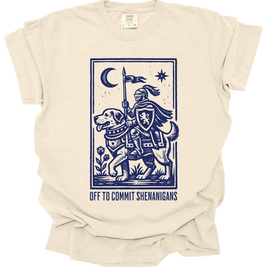 Ivory t-shirt. A knight in full armor rides a large hound like a horse, carrying a lance with a pennant, under a crescent moon and a star, all inside a thin ruled border. Underneath, "Off to commit shenanigans" in spaced capitals. Medieval woodcut style, all in one deep blue.