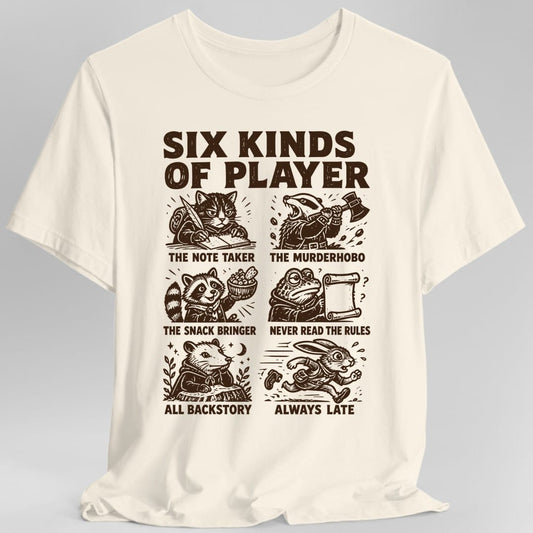 Natural t-shirt. The design reads SIX KINDS OF PLAYER in heavy dark brown capitals across the top, above a grid of six woodcut style animals, each labeled underneath. A cat writing with a quill, THE NOTE TAKER. A badger swinging an axe, THE MURDERHOBO. A raccoon holding a bowl of snacks, THE SNACK BRINGER. A frog squinting at a scroll, NEVER READ THE RULES. An opossum lounging, ALL BACKSTORY. A rabbit running, ALWAYS LATE.