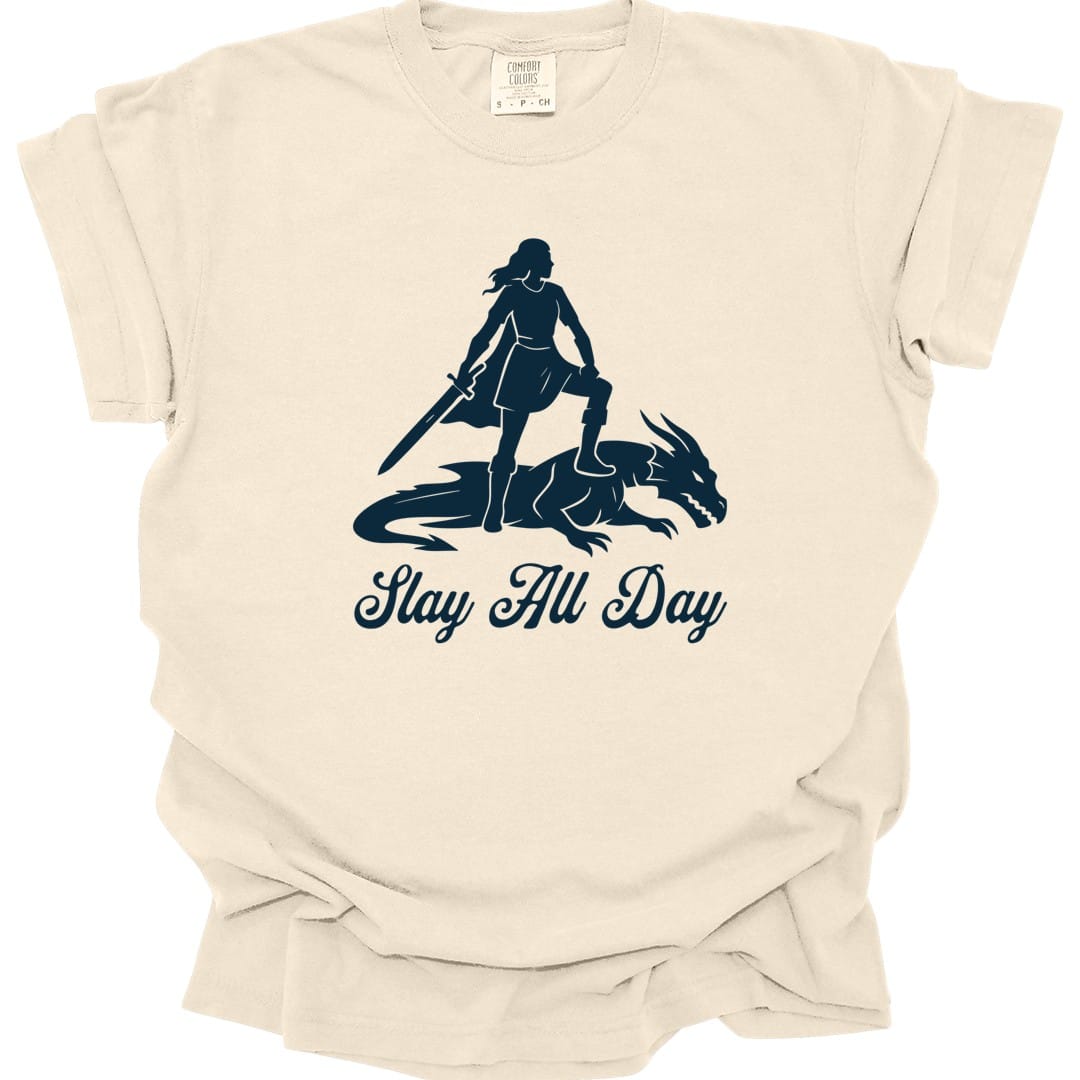 Ivory t-shirt. A woman stands in silhouette with one foot resting on the back of a slain dragon, holding a sword point down in front of her. Underneath, "Slay All Day" in a flowing script. All in a single deep navy blue.