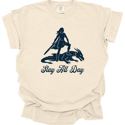Ivory t-shirt. A woman stands in silhouette with one foot resting on the back of a slain dragon, holding a sword point down in front of her. Underneath, "Slay All Day" in a flowing script. All in a single deep navy blue.